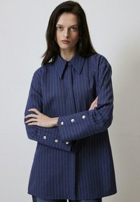 Navy blue striped shirt with a pointed collar, long sleeves featuring silver button accents, and a relaxed fit. Textured fabric with vertical lines.