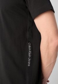 Čierne bavlnené tričko s krátkymi rukávmi, okrúhlym výstrihom a božnou etiketou s nápisom "Calvin Klein Jeans" v bielej výšivke.
