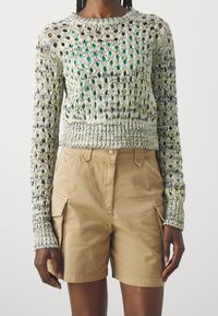 Osoba ubrana w zielono-kremowy sweter z długim rękawem i luźnym splotem na zielonej bluzce, w połączeniu z beżowymi szortami cargo do kolan.