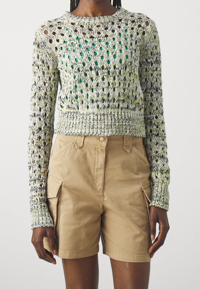 Osoba ubrana w zielono-kremowy sweter z długim rękawem i luźnym splotem na zielonej bluzce, w połączeniu z beżowymi szortami cargo do kolan.