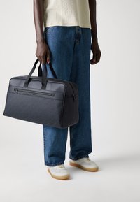 Black fabric duffle bag with textured front, dual top straps, and front zip pocket, held by a person wearing a light top and blue jeans.