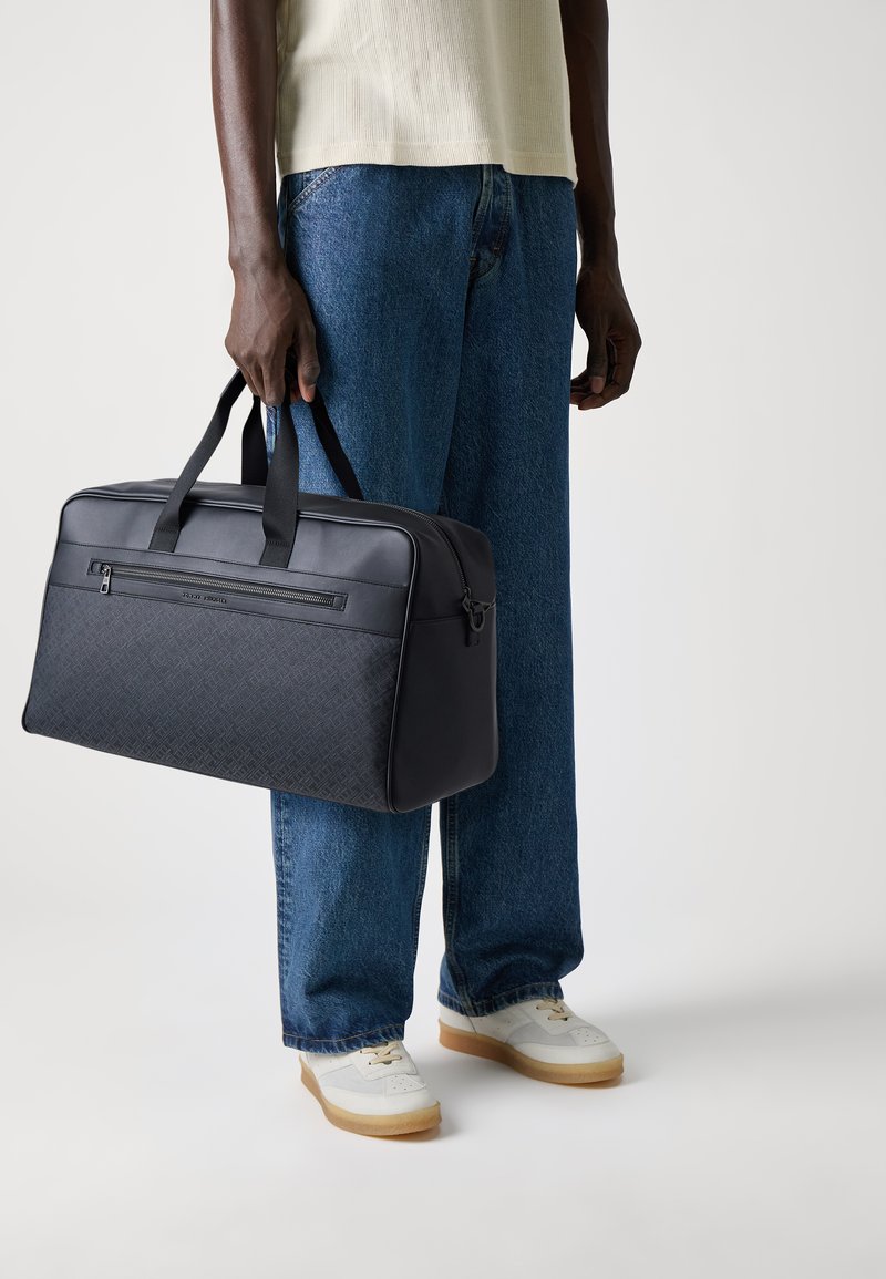 Black fabric duffle bag with textured front, dual top straps, and front zip pocket, held by a person wearing a light top and blue jeans.