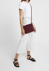 Burgundská semišová kabelka přes rameno s plochým designem a kontrastním páskem, spojená s bílými širokými kalhotami s proužky a černými sandály.