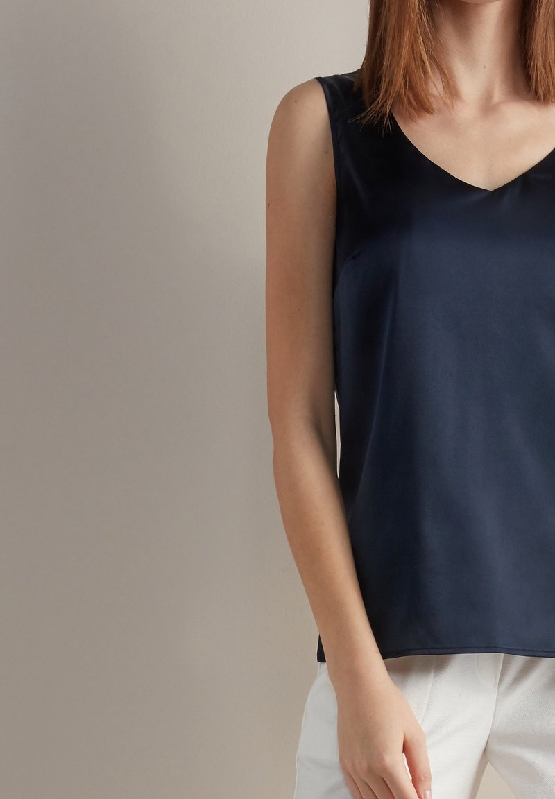 Femme portant un haut bleu marine sans manches et un pantalon blanc, debout devant un fond uni clair.