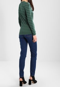 Grün gestreiftes Langarmshirt, getragen mit blauen Jeans und schwarzen offenen Absatzschuhen. Hat eine taillierte Silhouette und einen runden Ausschnitt.