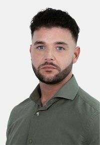 Een man met kort donker haar en een baard draagt een getailleerde groene shirt met knopen. Hij heeft blauwe ogen en een neutrale uitdrukking.