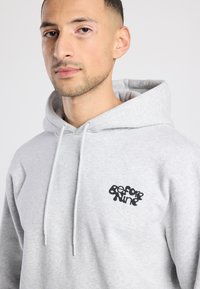 Grijze hoodie met een trekkoord, met een zwart grafisch logo op de linkerborst. Glad van textuur, losse pasvorm, effen kleur zonder patronen.