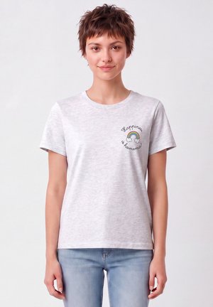 Femme aux cheveux courts portant un t-shirt gris clair avec un petit arc-en-ciel et le texte "Happiness is homemade" sur le côté gauche de la poitrine et un jean bleu clair.