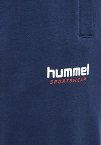 Mörkblå sportkläder i strukturerat tyg; har en vit och orange "hummel"-logotyp med "SPORTSWEAR" under i fet stil.