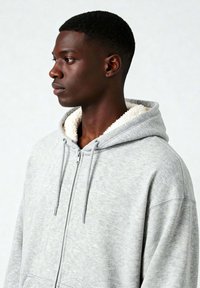 Felpa grigia con zip e un morbido rivestimento in pile crema sul cappuccio. Presenta lacci e una vestibilità rilassata. Tessuto testurizzato con un motivo sottile.