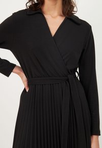 Černé zavinovací šaty s límečkem, dlouhými rukávy, páskem v pase a plisovanou sukní. Materiál působí měkce a hladce na dotek.