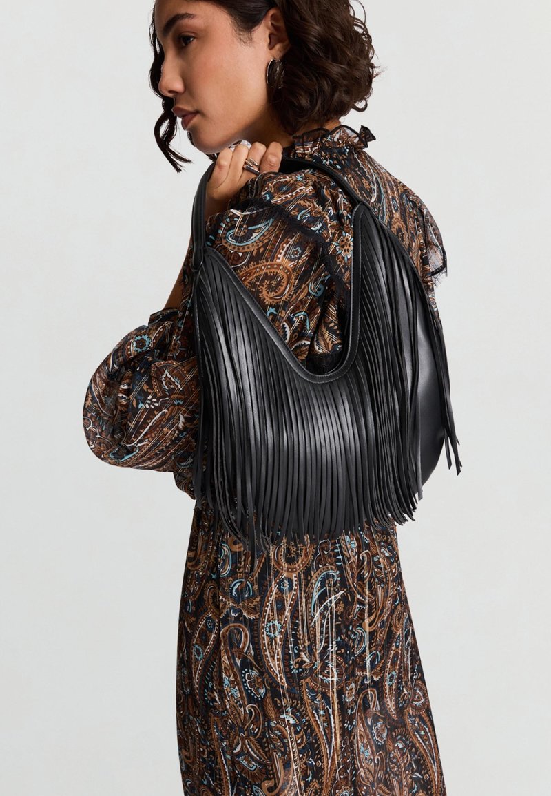 Borsa a tracolla in pelle nera con dettagli a frange lunghe, forma curva e texture liscia. Indossata sopra un vestito marrone con motivo.