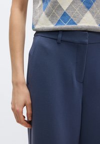 Παντελόνι φόρμας ναυτικού μπλε από υφασμάτινο ύφασμα με υφή, με στενή εφαρμογή, επίπεδη μπροστινή όψη και πλαϊνές τσέπες. Συνδυασμένο με γκρι πουλόβερ με σχέδιο.