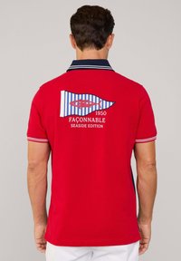 Camisa polo roja con cuello navy, que presenta un diseño de bandera a rayas azul y blanca y un motivo de pez. Incluye el texto "Fàçonable 1950 Edición de la Costa".