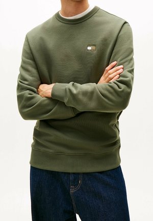 Person mit verschränkten Armen, die einen olivgrünen Tommy Jeans Pullover und blaue Jeans vor weißem Hintergrund trägt.