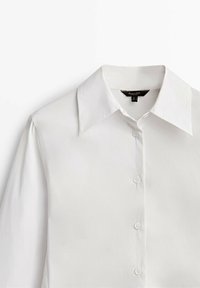 Chemise blanche à manches longues avec col pointu et boutons sur le devant, présentée à plat sur un fond blanc.