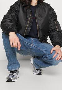 Svart bomberjacka med guld blixtlås, räfflade ärm- och nederkanter. Bärs över en marinblå skjorta och matchas med ljusblå denim och grå sneakers.