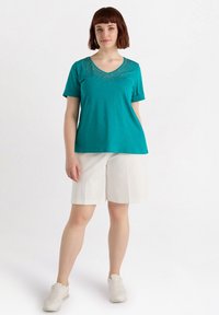 Top de manga corta en color azul verdoso con escote en V y destellos decorativos cerca del escote, combinado con pantalones cortos blancos y zapatillas blancas.