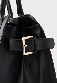 Czarna skórzana torebka o gładkiej fakturze, z akcentem w postaci złotego metalowego zapięcia i z pudełkowym fasonem. Uchwyty widoczne są na górze.