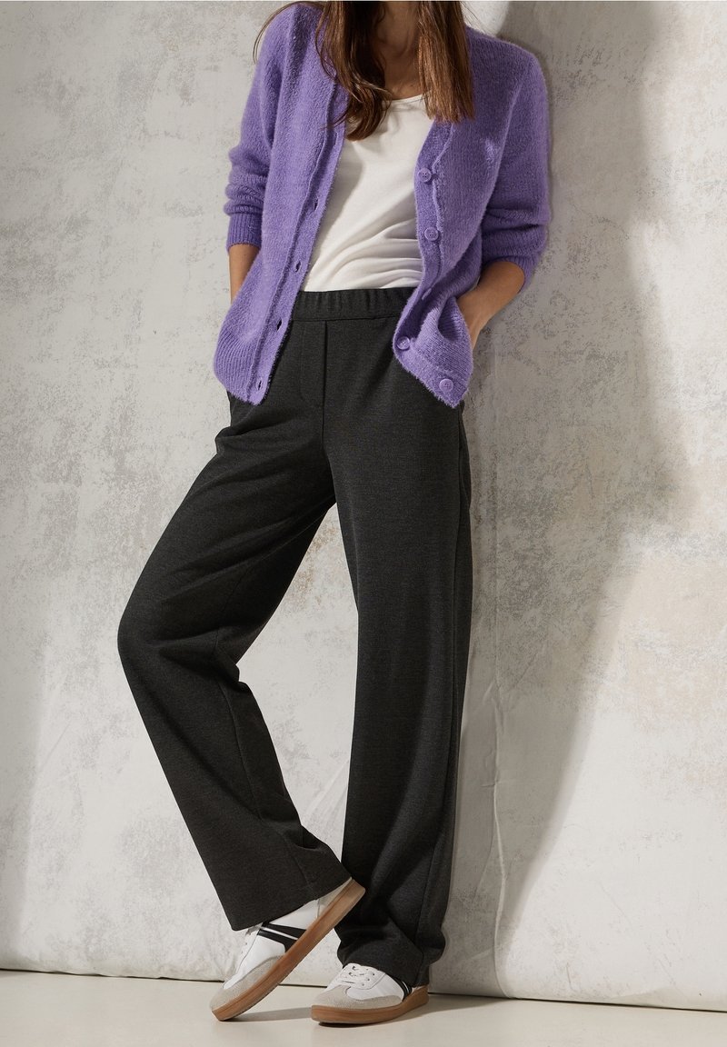 Femme portant un cardigan violet boutonné, un haut blanc, un pantalon large foncé et des baskets blanches, debout contre un mur clair texturé.