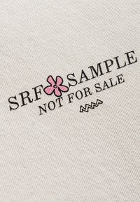Metka tekstylna z czarnym napisem "SRF SAMPLE" i różową grafiką kwiatu, umieszczona na jasnobeżowym, teksturowanym tle; zawiera napis "NIE NA SPRZEDAŻ".