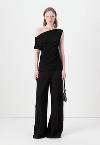 Melnas off-shoulder blūze ar savērptu faktūru, apvienota ar platām melnām biksēm ar kabatām. Modelis tur mazs melns soma.