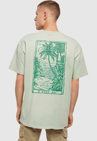 Mintgrøn bomulds T-shirt med et rygprint af palmer og en strandscene, omridset i mørkegrøn, med teksten "E KOMO MAI."