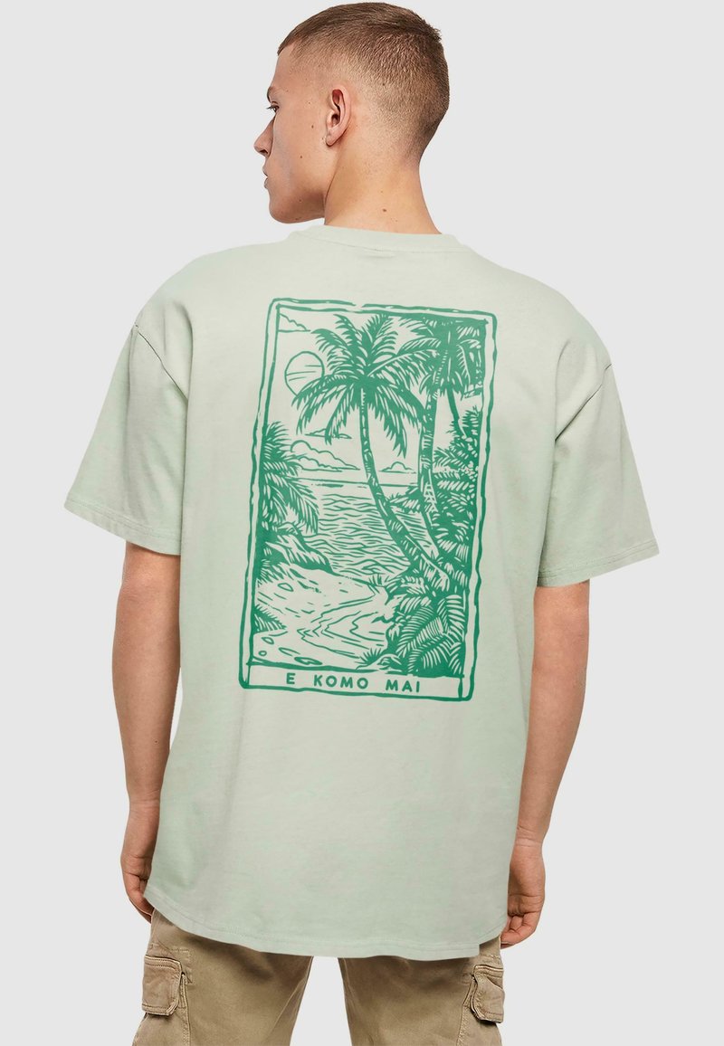 Minzgrünes Baumwoll-T-Shirt mit einem Grafik-Design auf der Rückseite, das Palmen und eine Strandlandschaft zeigt, umrandet von Dunkelgrün, mit dem Text "E KOMO MAI."