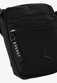 Svart axelväska gjord av slitstarkt tyg; har en ficka med dragkedja, präglad logotyp och en etikett med "JOARDAN" i vit text.