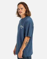 Man med lockigt hår som bär en blå T-shirt från DC Shoe Co. och visar en hajtatuering på sin vänstra underarm mot en enfärgad bakgrund.