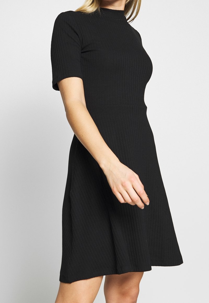 Femme portant une robe noire ajustée côtelée à manches courtes et col montant, debout devant un fond clair uni.