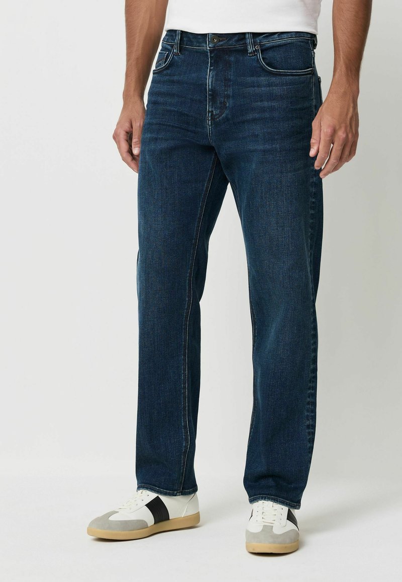 Dunkelblaue Jeans im geraden Schnitt mit gestickten Details, fünf Taschen und einem Metallknopfverschluss. Getragen mit weißen und schwarzen Turnschuhen.