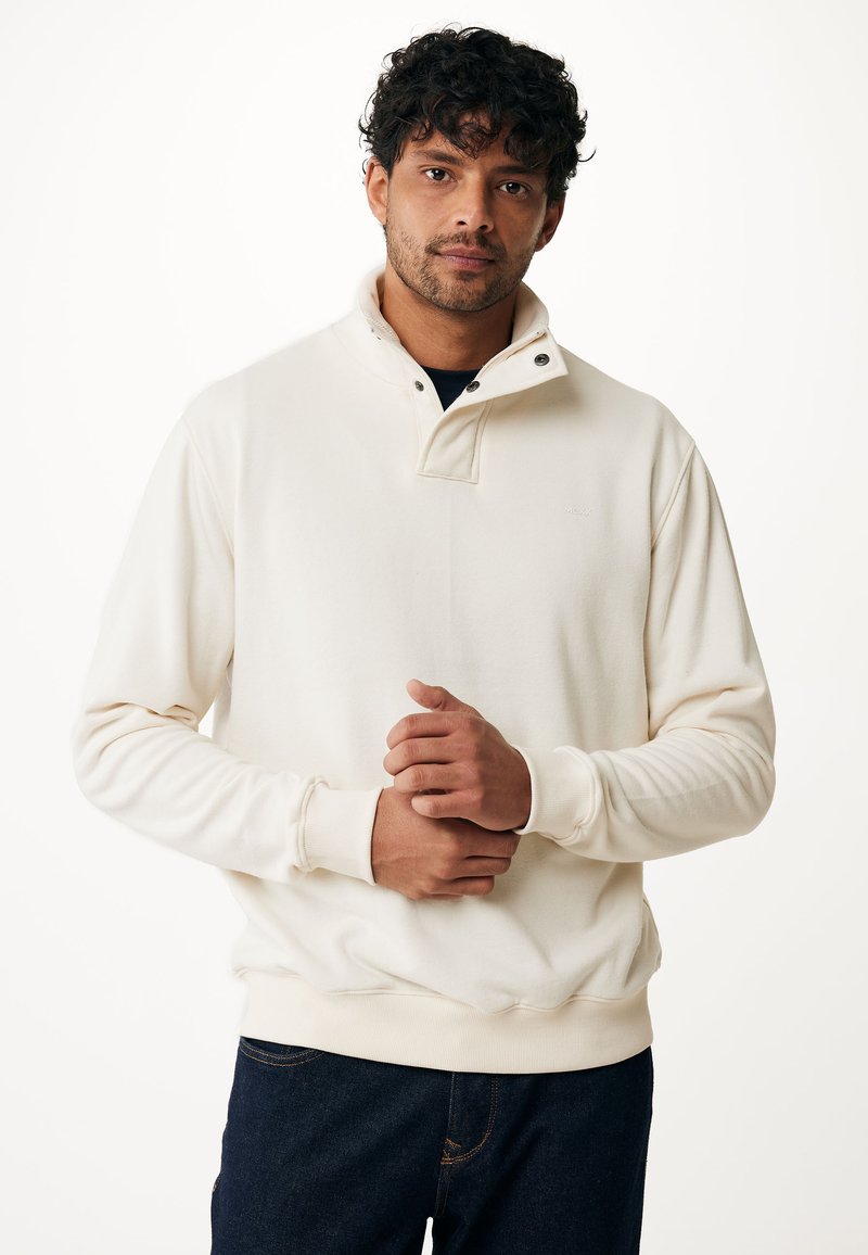 Sudadera color crema con un alto cuello y cierre a presión. La tela parece suave con puños de canalé, usada sobre una camiseta oscura.