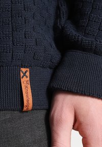 Marinblå stickad tröja med en strukturerad mönster, ribbade muddar och en brun läderetikett med logodetalj nära fållen.