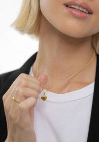 Arany szív medál nyaklánc vékony láncon, fehér ing fölött viselve. Arany gyűrű a kézen, amely a nyakláncot fogja, sima textúrával.