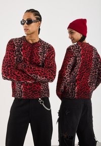 Suéter de leopardo en rojo y negro con mangas largas, textura de punto, diseño de cuello redondo, combinado con jeans negros desgastados y accesorios.