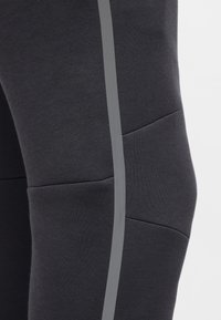 Melnas sporta bikses ar gludu auduma tekstūru, izceļoties ar izteiktu pelēku sānu svītru un izliekām šuvēm ceļā, veidojot skaidru formu.