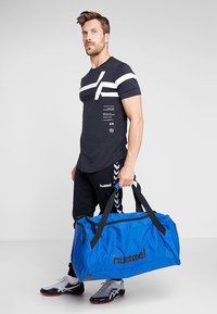 Bolsa de deporte azul con acentos negros, gran cremallera y logo. El modelo lleva una camiseta negra con rayas blancas y pantalones negros con logo.
