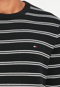 Maglione a righe bianche e nere in maglia, con un colletto a coste e un piccolo logo rosso, bianco e blu sul petto.