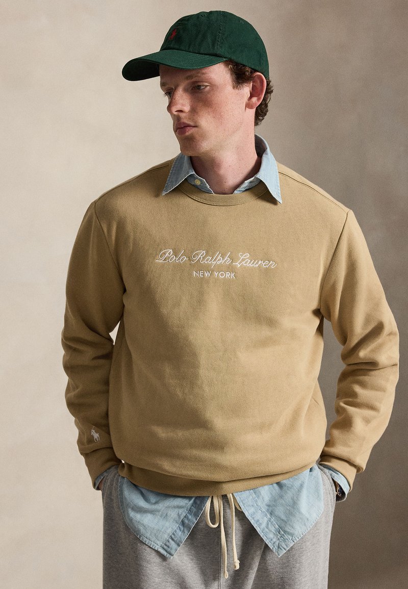 Νεαρός άνδρας φοράει μπεζ φούτερ Polo Ralph Lauren, ανοιχτό μπλε πουκάμισο, γκρι παντελόνι και σκούφο σκούρου πράσινου χρώματος, στέκεται με τα χέρια στις τσέπες.