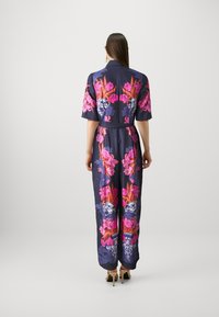 Blomstrete jumpsuit i marineblå sateng, med fargerike rosa og lilla blomster mønstre. Slim fit med belte og korte ermer. Høye hæler synlige.