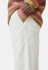Gebreide trui met rode, gele en bruine strepen; witte corduroy broek met zijzak en textuurstof. Hand rustend in de zak.