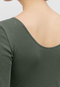 Grünes geripptes Oberteil mit tiefem Rückenausschnitt, figurbetontem Schnitt und weicher Textur, ausgestattet mit einem schlichten Halsausschnitt und langen Ärmeln.