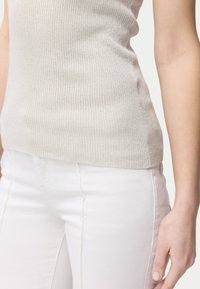 Närbild på en person som bär ett beige ribbat ärmlöst linne och figurnära vita byxor med en synlig sidoseam.