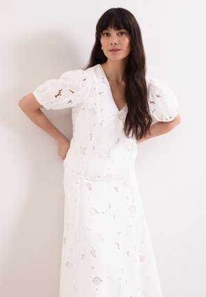 Donna con lunghi capelli scuri che indossa un top bianco in pizzo floreale con maniche a sbuffo e una gonna abbinata, in piedi con le mani sui fianchi su sfondo bianco.