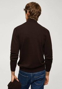 Bruine gebreide trui met een hoge kraag, nauwsluitende mouwen en een gladde textuur, gedragen over een blauwe spijkerbroek. Opvallend door het eenvoudige ontwerp.