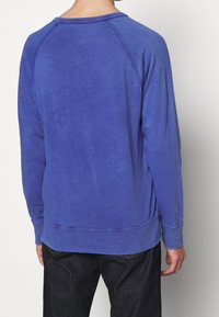 Sudadera de algodón azul con mangas largas, costuras raglán, puños acanalados y cuello redondo. Textura suave con un acabado ligeramente desvanecido.