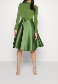 Top verde de manga larga con cuello con volantes, combinado con una falda de satén hasta la altura de la rodilla, ambos con un acabado brillante, acentuando una cintura ceñida.