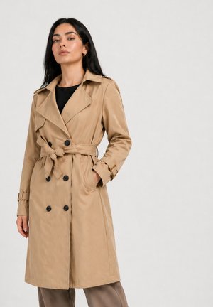 Kvinde iført en beige dobbeltradet trenchcoat bundet i taljen over en sort top, stående med den ene hånd i lommen mod en ensfarvet baggrund.