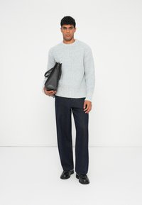 Maglione a coste grigio chiaro, pantaloni a gamba larga blu scuro e borsa in pelle nera, in piedi contro uno sfondo bianco uniforme.